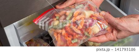A man reaches into the refrigerator and takes out a bag of frozen vegetables. He is preparing to cook during the afternoon in a kitchen setting, banner A man reaches into the refrigerator and takes out a bag of frozen vegetables. He is preparing to cook during the afternoon in a kitchen setting, banner 141793186