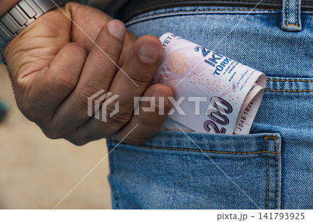 Close up view of man's hand of worker putting folded 200 Turkish lira banknotes into back pocket of his blue denim jeans 141793925