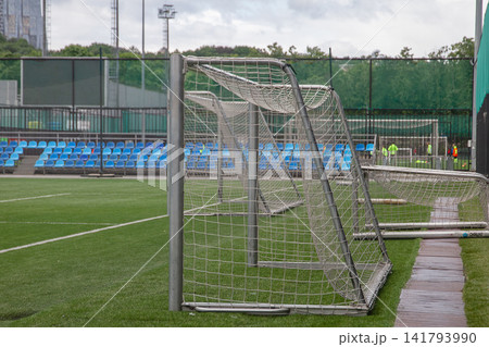 Close up view of metal training soccer goals with nets placed on side of green artificial grass football field with blue seats in background Close up view of metal training soccer goals with nets placed on side of green artificial grass football field with blue seats in background 141793990