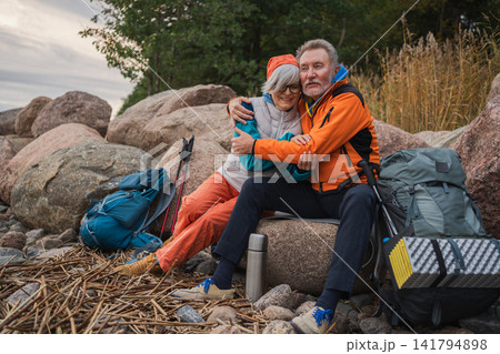 Hiking tourism adventure. Senior couple man woman traveling together enjoying outdoor recreation hiking having break sitting on beach. Happy old people backpackers hikers walking trekking on nature 141794898
