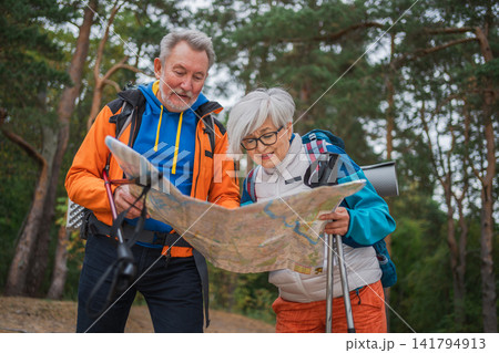 Hiking tourism. Senior couple man woman looking track in paper map enjoying hiking in forest. Happy old people backpackers hikers enjoy walking hike trekking tourism active vacation beauty of nature Hiking tourism. Senior couple man woman looking track in paper map enjoying hiking in forest. Happy old people backpackers hikers enjoy walking hike trekking tourism active vacation beauty of nature 141794913