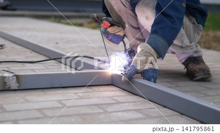 Close view of manual metal welding on ground level frame outdoors, showing bright arc light, protective gloves, worn work clothing, and precise fabrication work on construction site 141795861