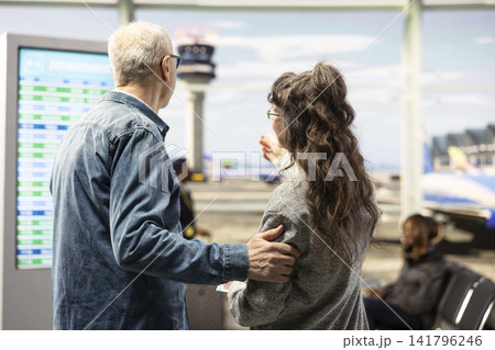 Joyful couple, checking flight status, and boarding gate information, elderly man and woman standing near airport screen in terminal concourse. Senior couple using screen for wayfinding. Joyful couple, checking flight status, and boarding gate information, elderly man and woman standing near airport screen in terminal concourse. Senior couple using screen for wayfinding. 141796246