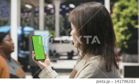 Green screen smartphone used by woman walking inside bus terminal. Female passenger looking at chroma key phone display, waiting for shuttle in coach station 141796283