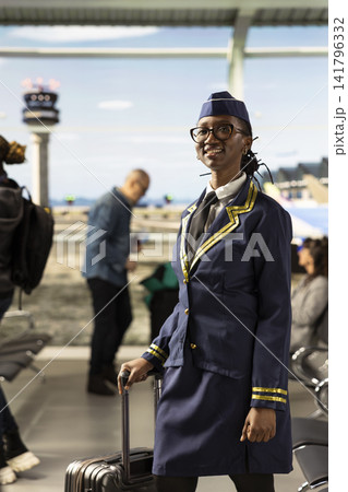 Smiling black flight attendant in uniform walking through busy airport terminal with rolling suitcase, ready for departure. Cabin crew member representing airline service in travel industry. Smiling black flight attendant in uniform walking through busy airport terminal with rolling suitcase, ready for departure. Cabin crew member representing airline service in travel industry. 141796332