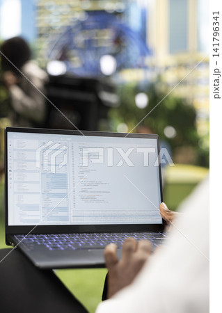 Close up of person working with programming interface and source code on laptop, representing software development and flexible IT work in city park. Over-the-shoulder view. 141796341