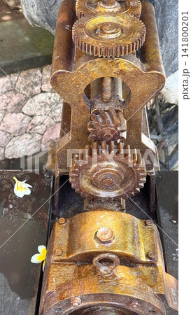 Rusty interlocking metal gears on vintage mechanical winch with textured corrosion, worn teeth, warm patina, threaded shaft, and heavy aged surface beside stone paving Rusty interlocking metal gears on vintage mechanical winch with textured corrosion, worn teeth, warm patina, threaded shaft, and heavy aged surface beside stone paving 141800201