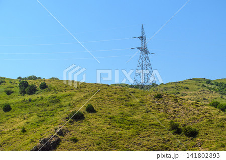 Electricity pylon rises above rolling green hills under bright blue sky Electricity pylon rises above rolling green hills under bright blue sky 141802893