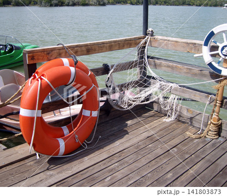 Wooden pier corner with an orange lifebuoy, a draped fishing net adorned with a skull, and a decorative ship's wheel. A calm lake and boats are in the background under a sunny sky Wooden pier corner with an orange lifebuoy, a draped fishing net adorned with a skull, and a decorative ship's wheel. A calm lake and boats are in the background under a sunny sky 141803873