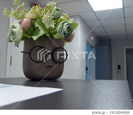 A quirky face-shaped planter wearing round glasses sits on a dark desk. It holds a bouquet of faux teal and pink flowers, positioned against a background of a modern office corridor A quirky face-shaped planter wearing round glasses sits on a dark desk. It holds a bouquet of faux teal and pink flowers, positioned against a background of a modern office corridor 141804086