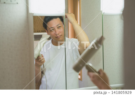 洗面台の鏡に映る笑顔の40代男性がドライヤーで髪を乾かすヘアケア 洗面台の鏡に映る笑顔の40代男性がドライヤーで髪を乾かすヘアケア 141805929