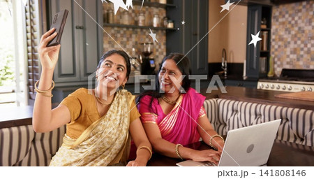 Smiling daughter holding smartphone, mother watching laptop at kitchen nook in sarees 141808146