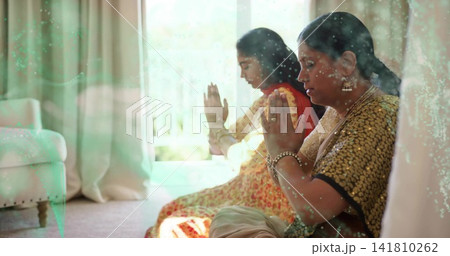 Praying Indian mother and daughter sitting in living room by window, wearing sari, dupatta, jewelry Praying Indian mother and daughter sitting in living room by window, wearing sari, dupatta, jewelry 141810262