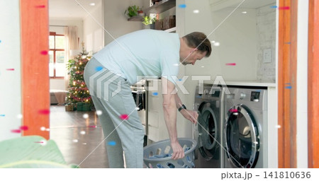 Bending man in blue tee, sweatpants loading washer in laundry nook, holding basket, Christmas tree Bending man in blue tee, sweatpants loading washer in laundry nook, holding basket, Christmas tree 141810636