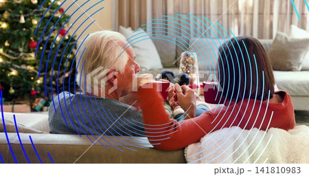 Sipping senior couple wearing sweaters, sitting on sofa at home with blue overlay, holding red mugs 141810983