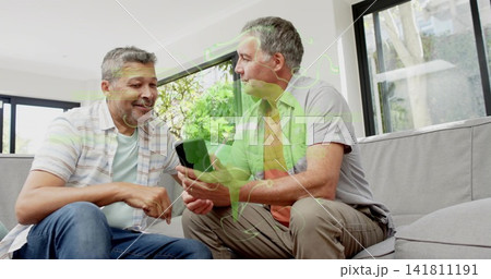 Smiling mature men wearing casual shirts, pants, viewing black smartphone on grey couch at home 141811191
