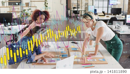 Leaning three women in casual attire pointing at wood table and sticky notes, showing data overlays 141811890