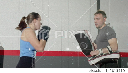 Throwing punches female trainee in blue tank, black gloves, male coach holding Thai pads in gym 141813410