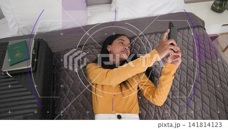 Lying woman in mustard top light pants holding smartphone on bed, with suitcase, passport and HUD Lying woman in mustard top light pants holding smartphone on bed, with suitcase, passport and HUD 141814123