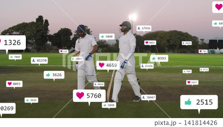 Walking two cricket players wearing helmets, pads holding bats on dusk field, with social icons 141814426