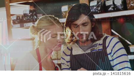 Inspecting two staff in aprons checking tablet at shop with wood shelves and jars, data overlay 141815616