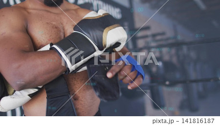 Adjusting shirtless boxer securing black strap at boxing gym near ring, with blue wraps and glove 141816187