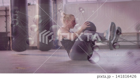 Twisting mid-adult woman holding medicine ball in gym by punching bags, in dark tank top, overlay Twisting mid-adult woman holding medicine ball in gym by punching bags, in dark tank top, overlay 141816426
