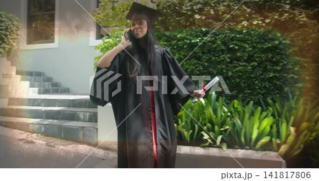 Talking graduate wearing black gown and cap, standing on campus steps holding diploma and phone Talking graduate wearing black gown and cap, standing on campus steps holding diploma and phone 141817806