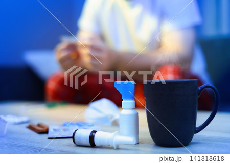 Cold and flu medicine, throat spray, pills, and a hot drink on a table. Blurred sick man resting in the background, recovering from seasonal respiratory illness at home. Cold and flu medicine, throat spray, pills, and a hot drink on a table. Blurred sick man resting in the background, recovering from seasonal respiratory illness at home. 141818618