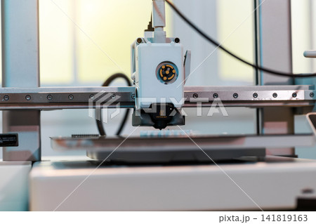 Close up of a 3d printer head working on a build plate in a workshop, with blurred cables and machine parts in the background. 141819163