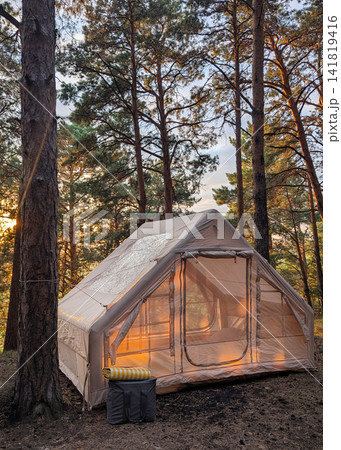 Glamping tent glowing at sunset among tall pine trees inside quiet forest campsite, warm interior light, soft sky behind branches, rustic outdoor retreat in nature Glamping tent glowing at sunset among tall pine trees inside quiet forest campsite, warm interior light, soft sky behind branches, rustic outdoor retreat in nature 141819416