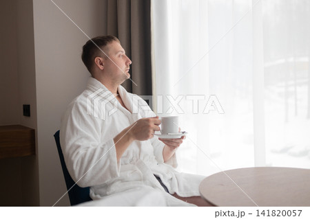 Young man in white bathrobe drinking coffee by window in hotel room 141820057