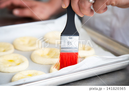 Close up hand brushing pastry dough on tray with red bristles in bakery kitchen, preparing round rolls before baking with soft focus background and stainless steel workspace Close up hand brushing pastry dough on tray with red bristles in bakery kitchen, preparing round rolls before baking with soft focus background and stainless steel workspace 141820365
