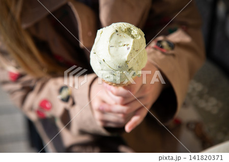 person holding ice cream cone in casual indoor setting with trench coat and green ice cream, close-up shot from below the cone, possibly a cafe or store, green and brown tones, casual. person holding ice cream cone in casual indoor setting with trench coat and green ice cream, close-up shot from below the cone, possibly a cafe or store, green and brown tones, casual. 141820371