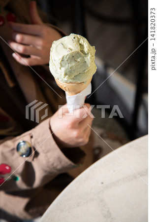 person in trench coat holding ice cream cone in casual outdoor seating area above close-up view of wax paper and wooden cone on table with green and brown tones. person in trench coat holding ice cream cone in casual outdoor seating area above close-up view of wax paper and wooden cone on table with green and brown tones. 141820373