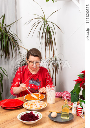 In a cozy home setting, a woman mixes layers of boiled vegetables with mayonnaise to prepare Olivier salad. The scene shows holiday decorations and festive plants nearby In a cozy home setting, a woman mixes layers of boiled vegetables with mayonnaise to prepare Olivier salad. The scene shows holiday decorations and festive plants nearby 141820628