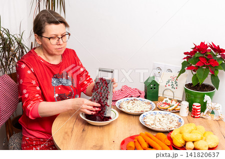 A woman grates beets for the Shuba salad on a Christmas table. Various ingredients are laid out, including herring, potatoes, eggs, and carrots, creating a festive atmosphere for the celebration A woman grates beets for the Shuba salad on a Christmas table. Various ingredients are laid out, including herring, potatoes, eggs, and carrots, creating a festive atmosphere for the celebration 141820637