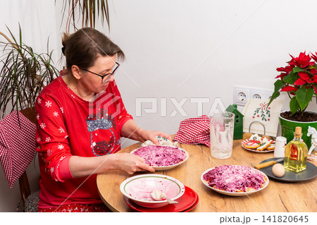 Woman works on preparing Olivier salad for Christmas. She carefully arranges layers of boiled vegetables, adding mayonnaise in a cozy kitchen setting surrounded by plants Woman works on preparing Olivier salad for Christmas. She carefully arranges layers of boiled vegetables, adding mayonnaise in a cozy kitchen setting surrounded by plants 141820645