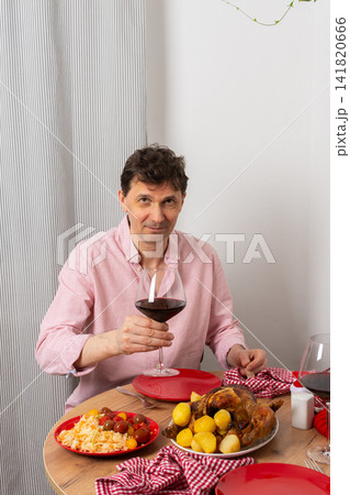 A man is cooking dinner for Christmas by baking chicken with potatoes in his kitchen. He is sitting at the table, ready to enjoy the meal with a glass of wine 141820666