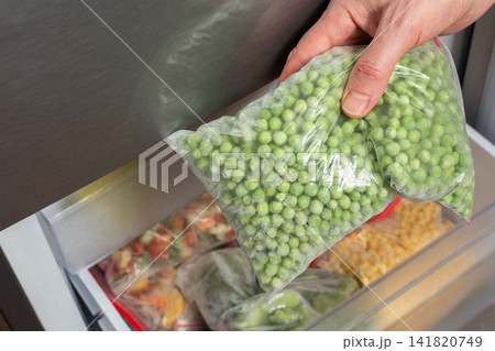 A man opens the refrigerator to take out a bag of frozen green peas. He is in the kitchen, preparing food for a meal in the afternoon. The refrigerator is organized with several bags inside A man opens the refrigerator to take out a bag of frozen green peas. He is in the kitchen, preparing food for a meal in the afternoon. The refrigerator is organized with several bags inside 141820749