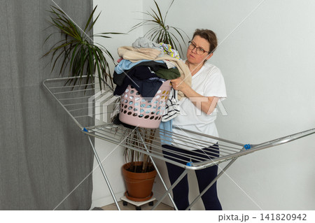 A woman is taking laundry out of the dryer and putting it into a basket. She is in a simple room with plants nearby. Her focus is on the task at hand A woman is taking laundry out of the dryer and putting it into a basket. She is in a simple room with plants nearby. Her focus is on the task at hand 141820942