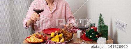 A man is cooking dinner for Christmas by baking chicken with potatoes in his kitchen. He is sitting at the table, ready to enjoy the meal with a glass of wine, banner 141820969