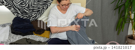 A 52-year-old woman is sorting clean laundry on a bed. She holds a pair of pants in one hand while looking at the clothes around her. A plant is seen in the corner of the room, banner A 52-year-old woman is sorting clean laundry on a bed. She holds a pair of pants in one hand while looking at the clothes around her. A plant is seen in the corner of the room, banner 141821017