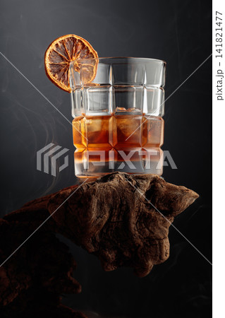 Smoked old-fashioned cocktail with ice and a dried orange slice served on weathered driftwood. Smoked old-fashioned cocktail with ice and a dried orange slice served on weathered driftwood. 141821477