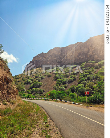 Scenic mountain road winds through sunlit hills with green vegetation 141821554