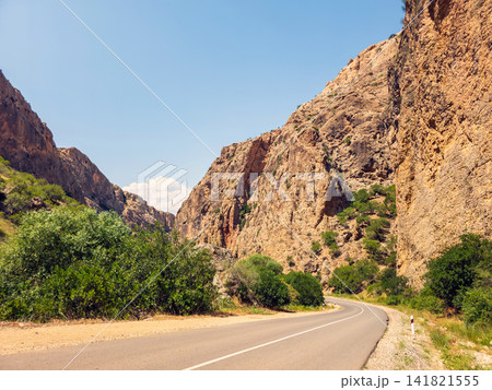 Road curves through arid canyon with towering rock faces and greenery Road curves through arid canyon with towering rock faces and greenery 141821555