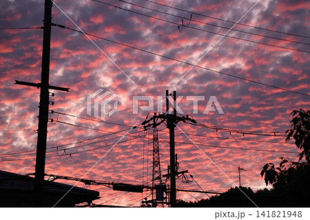 茜色の夕焼け空とシルエットの電柱や電線と樹々と家の屋根の風景 141821948