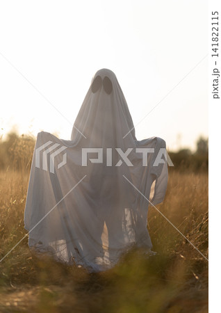 Backlit child in ghost costume holding sheet outdoors at sunset 141822115
