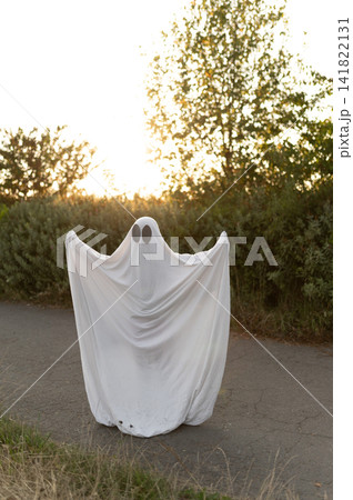 Halloween ghost standing on asphalt path near bushes at sunset Halloween ghost standing on asphalt path near bushes at sunset 141822131