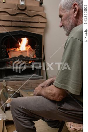 Old man sitting alone near home fireplace. Senior lifestyle. Old tired man watching flame in fireplace. Sad grey man burning logs and paper. Cozy winter evening. Alternative heat. Old man sitting alone near home fireplace. Senior lifestyle. Old tired man watching flame in fireplace. Sad grey man burning logs and paper. Cozy winter evening. Alternative heat. 141823803